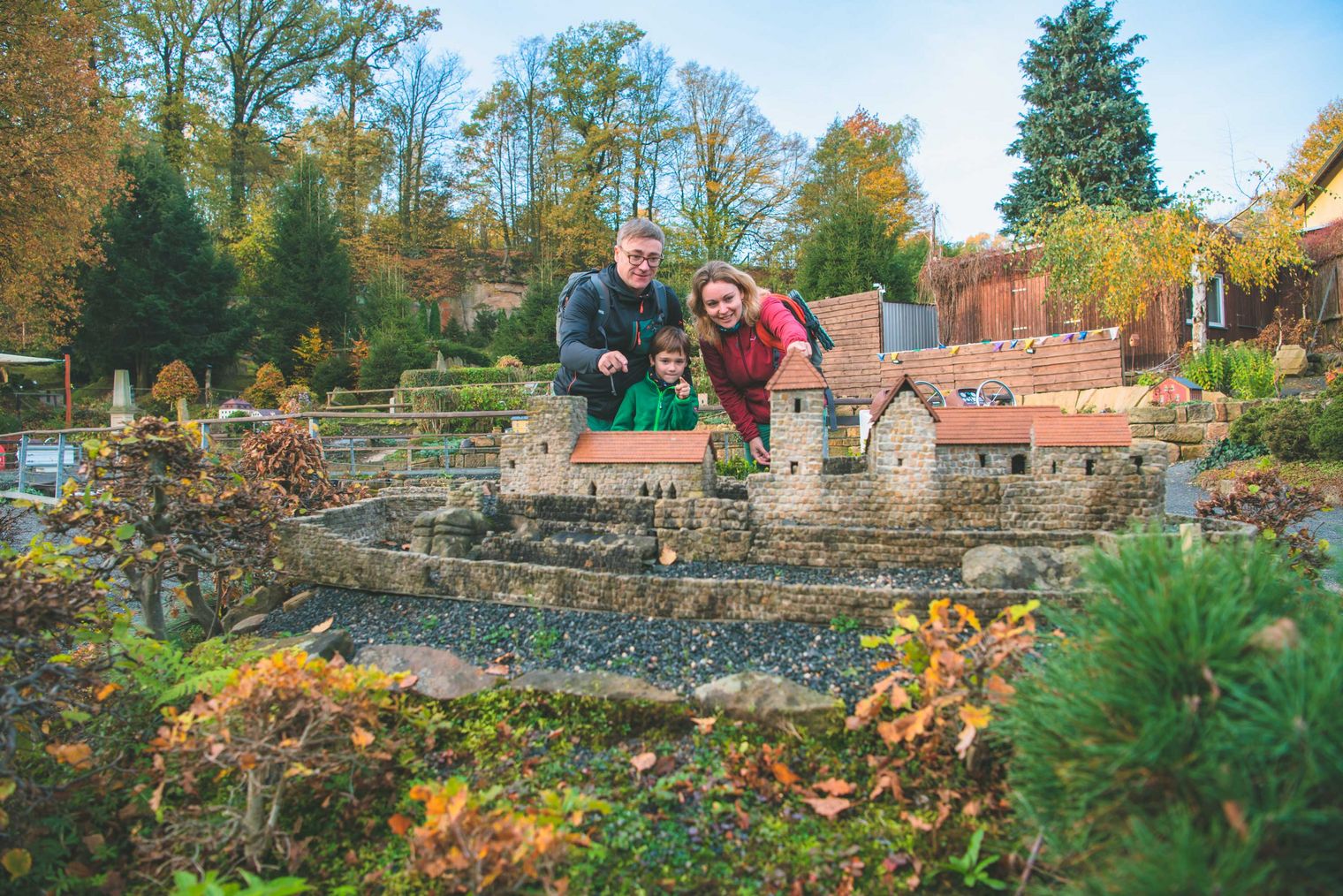 Paar mit Kind posiert vor einem Miniaturmodell eines Hauses mit Garten und Wasserlauf. Herbstliche Bäume im Hintergrund.