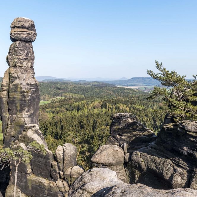 Felsnadel Barbarine am Fels Pfaffenstein und Blick auf eine grüne Landschaft unter blauem Himmel.