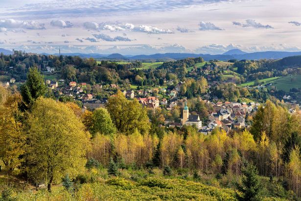 Blick auf Sebnitz: Häuser und Kirchturm umgeben von bewaldeten Hügeln unter blauem Himmel.