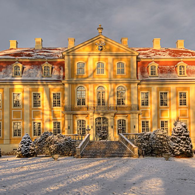 Historisches Gebäude mit gelber Fassade, Schnee auf dem Boden und Bäumen, im warmen Licht der Abenddämmerung.