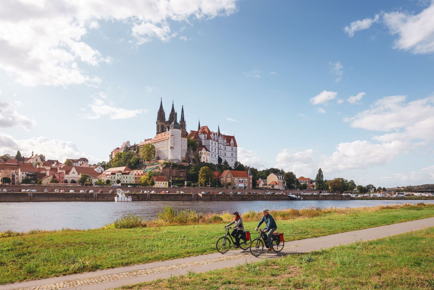 Zwei Radfahrer fahren entlang eines Flusses mit einer Burg im Hintergrund und blauem Himmel.