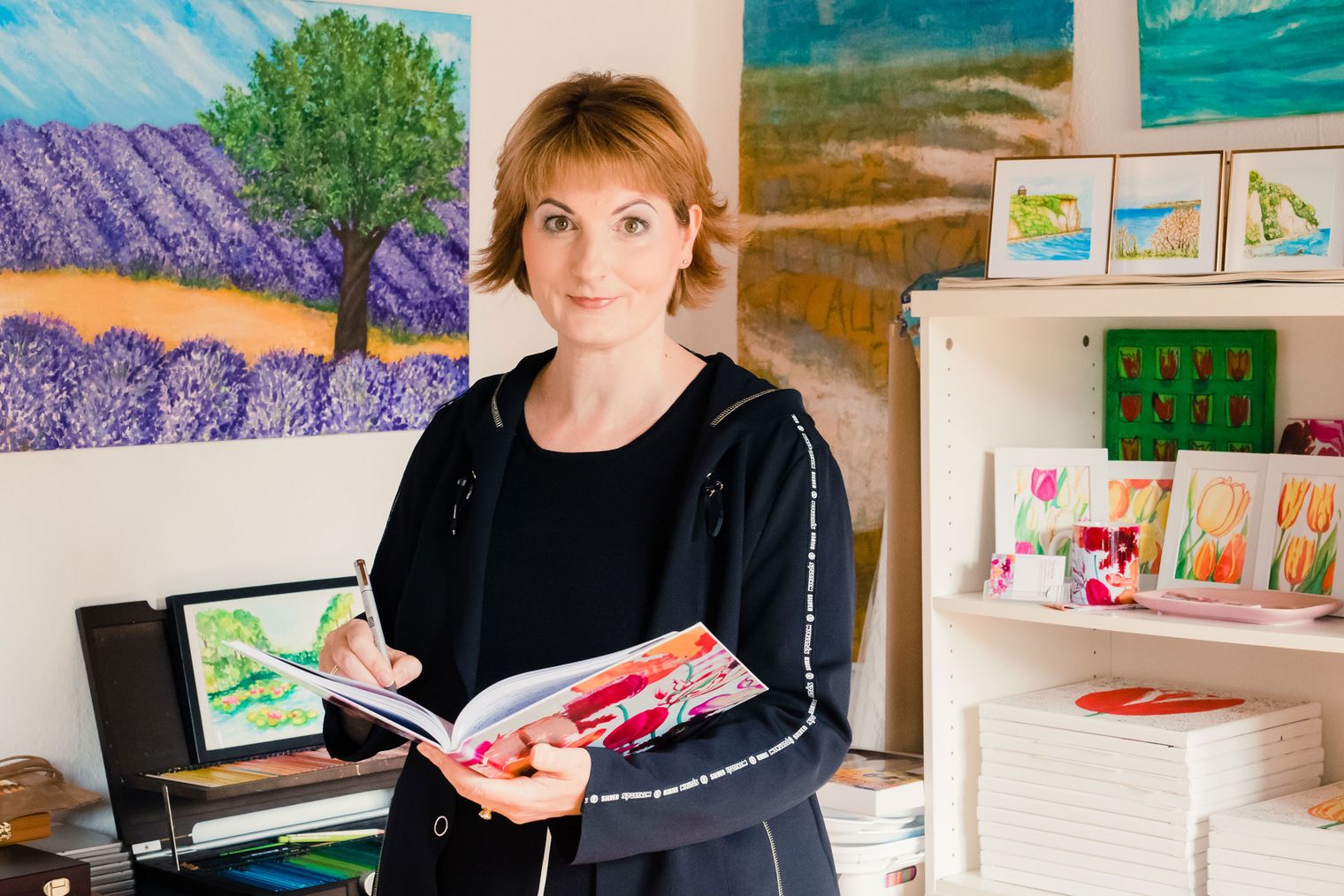 Künstlerin mit kurzem Haar hält ein Buch in einem Raum mit bunten Bildern und Pflanzen.