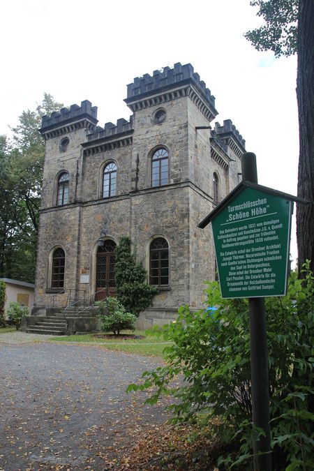 Historisches Schloss mit Türmen, umgeben von Bäumen und einem Weg. Ein grünes Schild steht im Vordergrund.