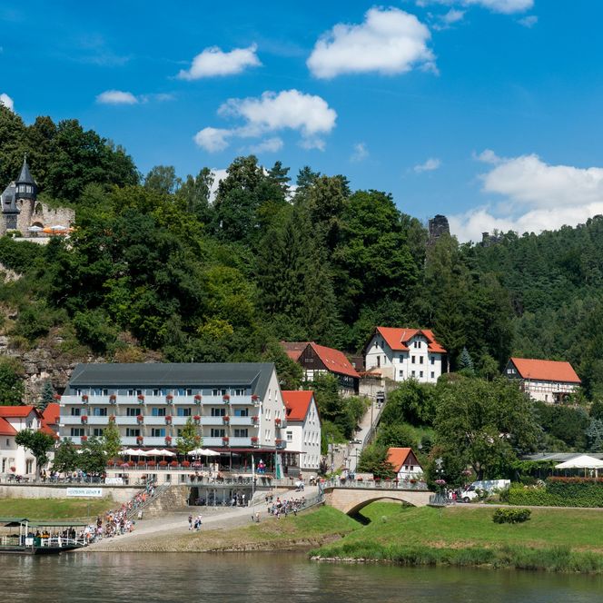 Blick auf ein malerisches Ufer mit einem Hotel, Häusern und bewaldeten Hügeln unter blauem Himmel.