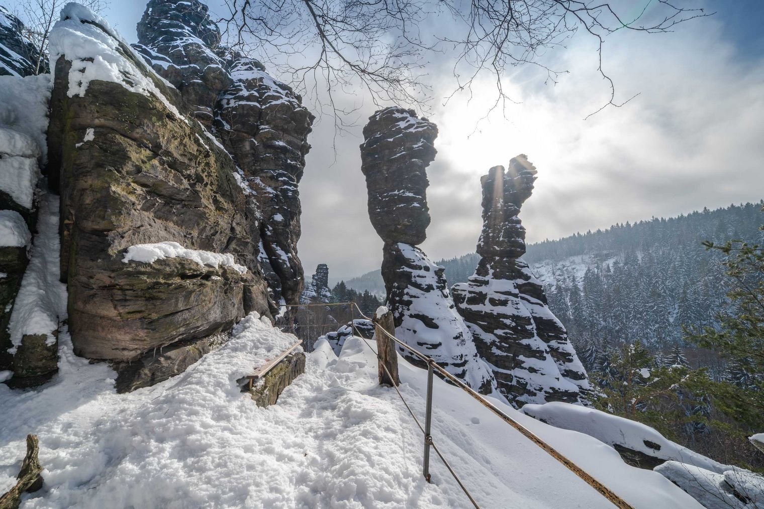 Schneebedeckte Felsenlandschaft im Bielatal mit Sonnenstrahlen und bewölktem Himmel im Hintergrund.
