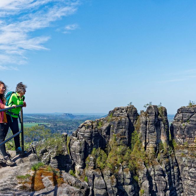 Zwei Wanderer stehen auf einem Felsen und blicken auf die Landschaft an der Schrammsteinaussicht unter blauem Himmel.