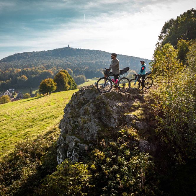 Zwei Mountainbiker stehen auf einem Felsen mit Blick auf eine hügelige Landschaft und einen Fernsehturm im Hintergrund.