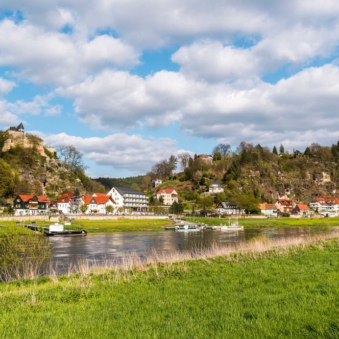 Idyllische Landschaft mit einem Fluss, Schiff, Fähre, einem Dorf und bewaldeten Hügeln unter einem bewölkten Himmel.