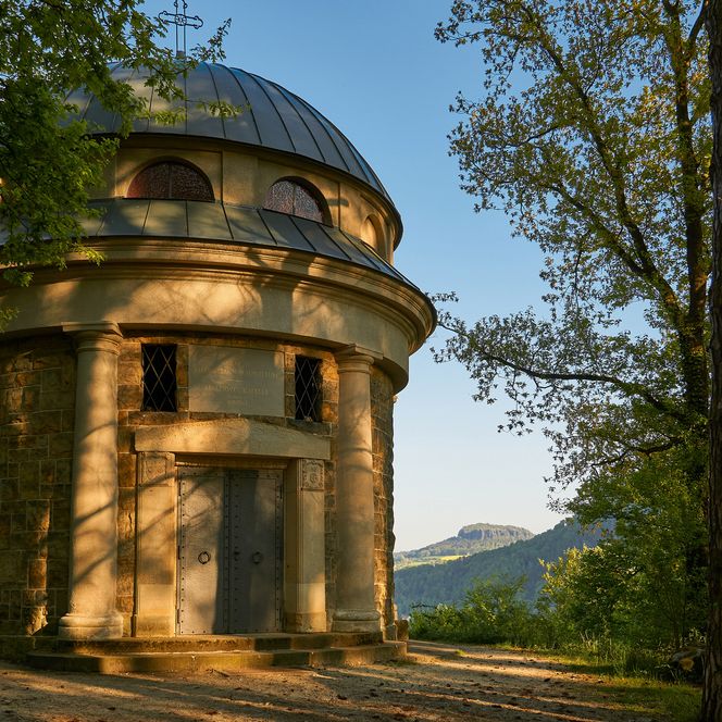 Die historische Malerwegkapelle umgeben von Bäumen und sanften Hügeln im Hintergrund.