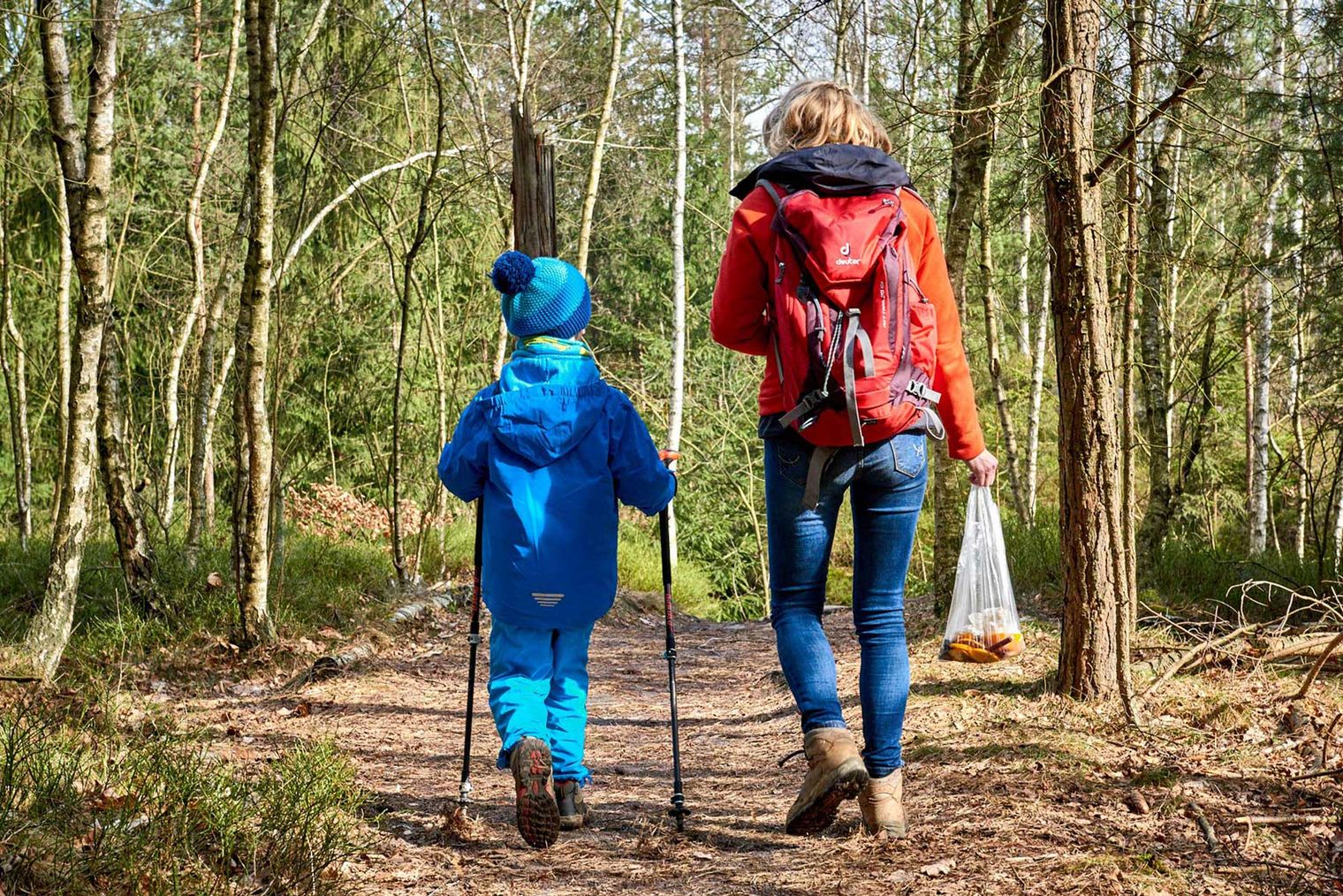 Eine Frau in roter Jacke und ein Kind in blauer Kleidung gehen mit Wanderstöcken einen Waldweg entlang.