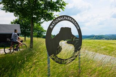 Ein Radfahrer steht neben einem Schild mit der Aufschrift "Nationalparkregion Sächsische Schweiz" vor einer grünen Landschaft.