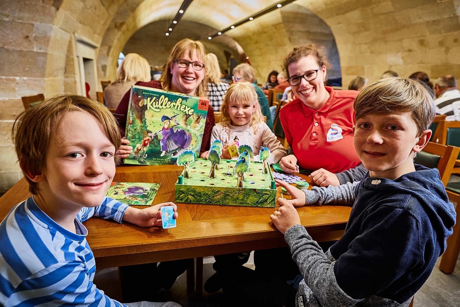 Fünf Kinder und Erwachsene sitzen an einem Tisch mit einem Brettspiel und lächeln in die Kamera.