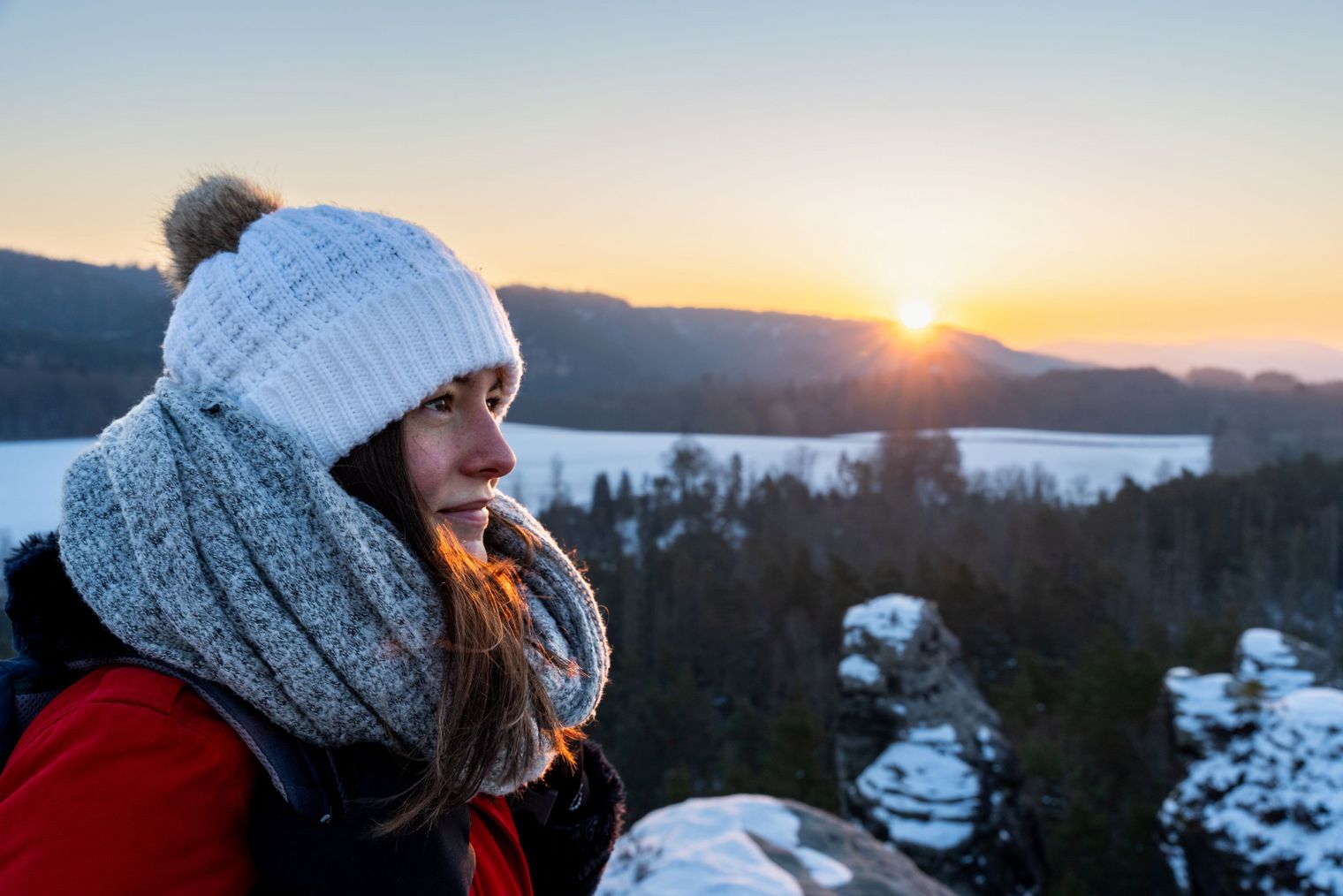 Frau mit Mütze und Schal blickt lächelnd auf einen Sonnenuntergang über einer winterlichen Landschaft.