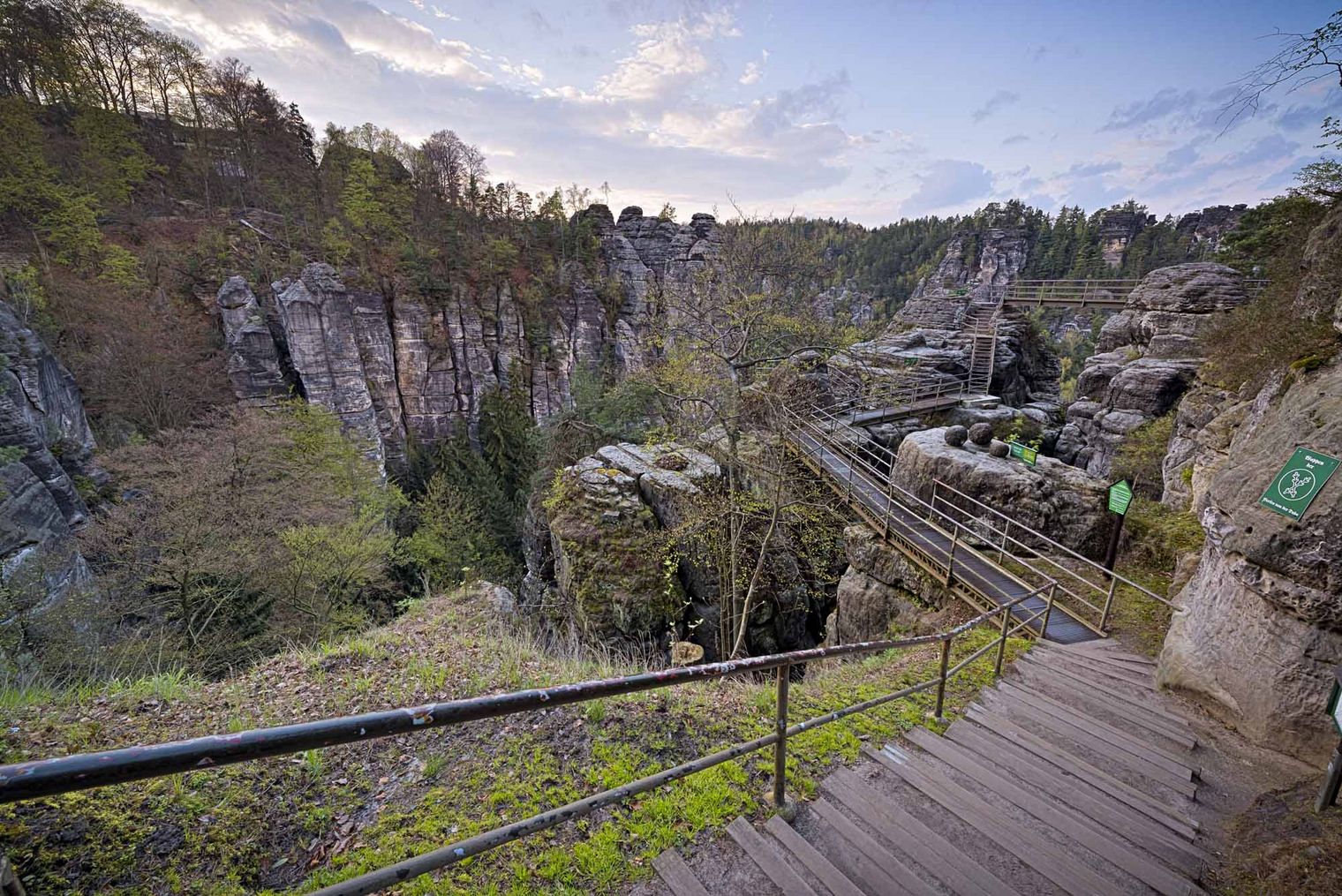 Holztreppe führt durch eine felsige Schlucht mit Bäumen und Felsen im Hintergrund.