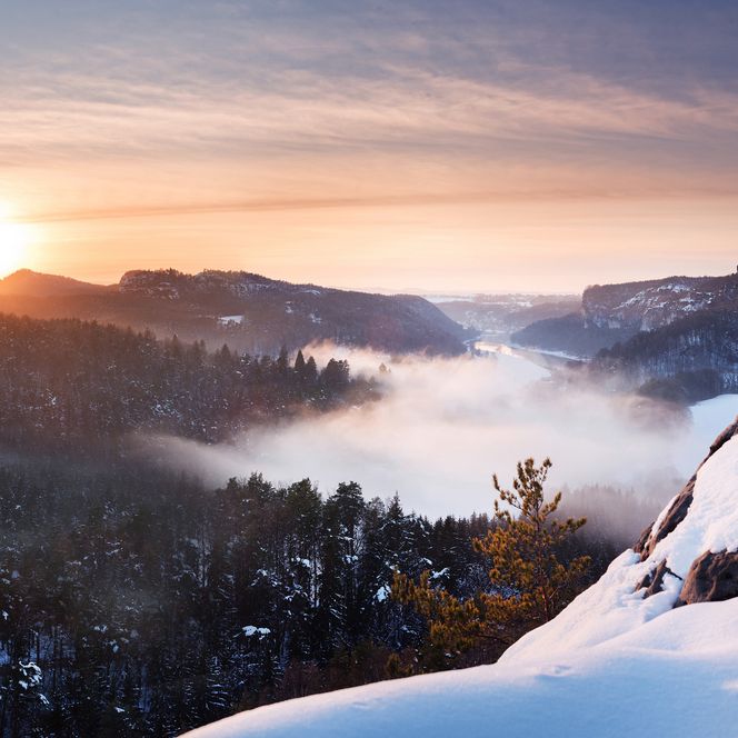Sonnenaufgang über einer verschneiten Landschaft mit Nebel im Tal und bewaldeten Hügeln.