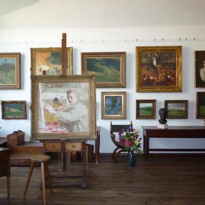 Atelier mit mehreren gerahmten Gemälden an der Wand, einem Malpult und einem Holzstuhl im Vordergrund.