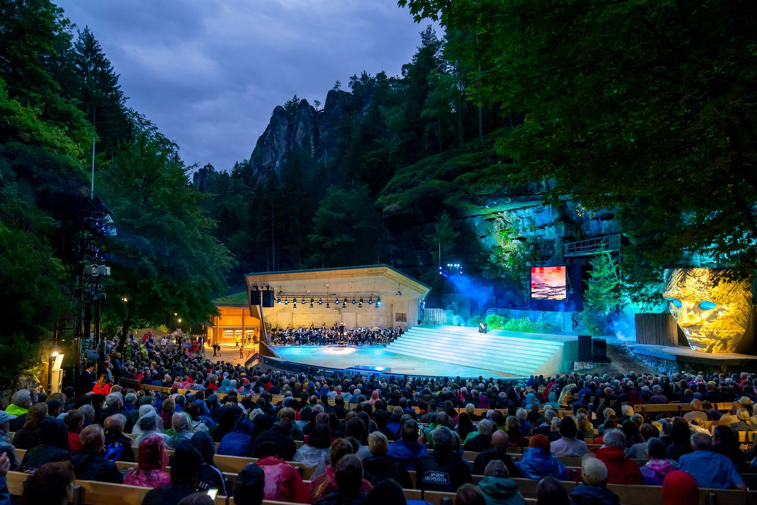 Die Felsenbühne in einer natürlichen Umgebung eingebettet, umgeben von Bäumen, mit vielen Zuschauern und bunten Lichtern.