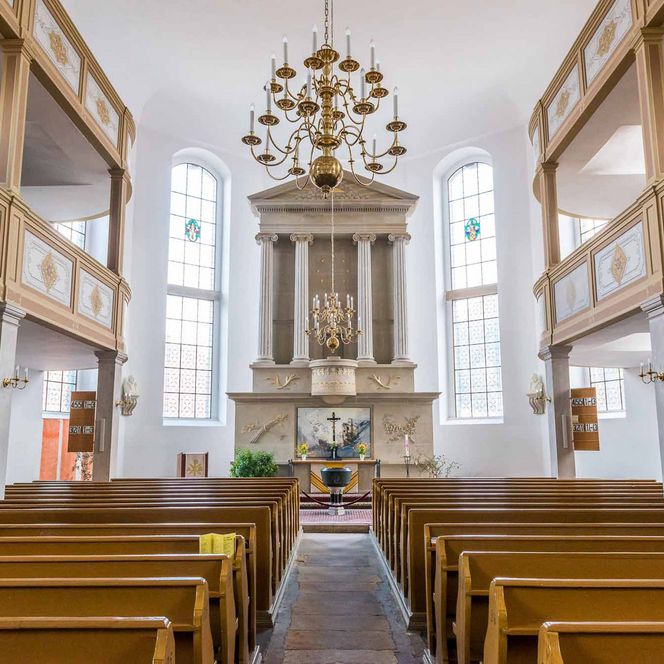Innenansicht einer Kirche mit Holzbankreihen, einem Altar und großen Fenstern.