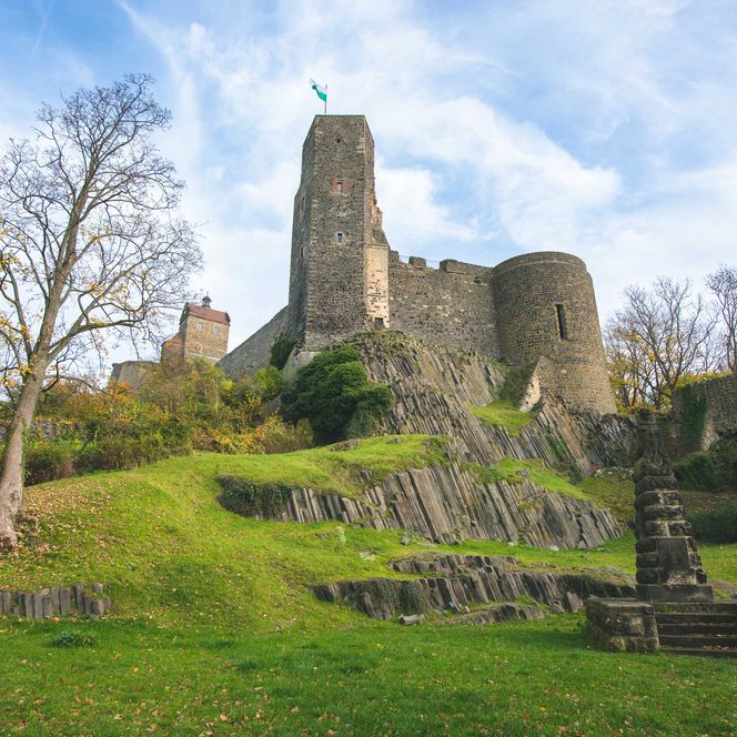Historische Burg Stolpen auf einem Hügel, umgeben von Bäumen und grünem Gras, unter einem blauen Himmel.
