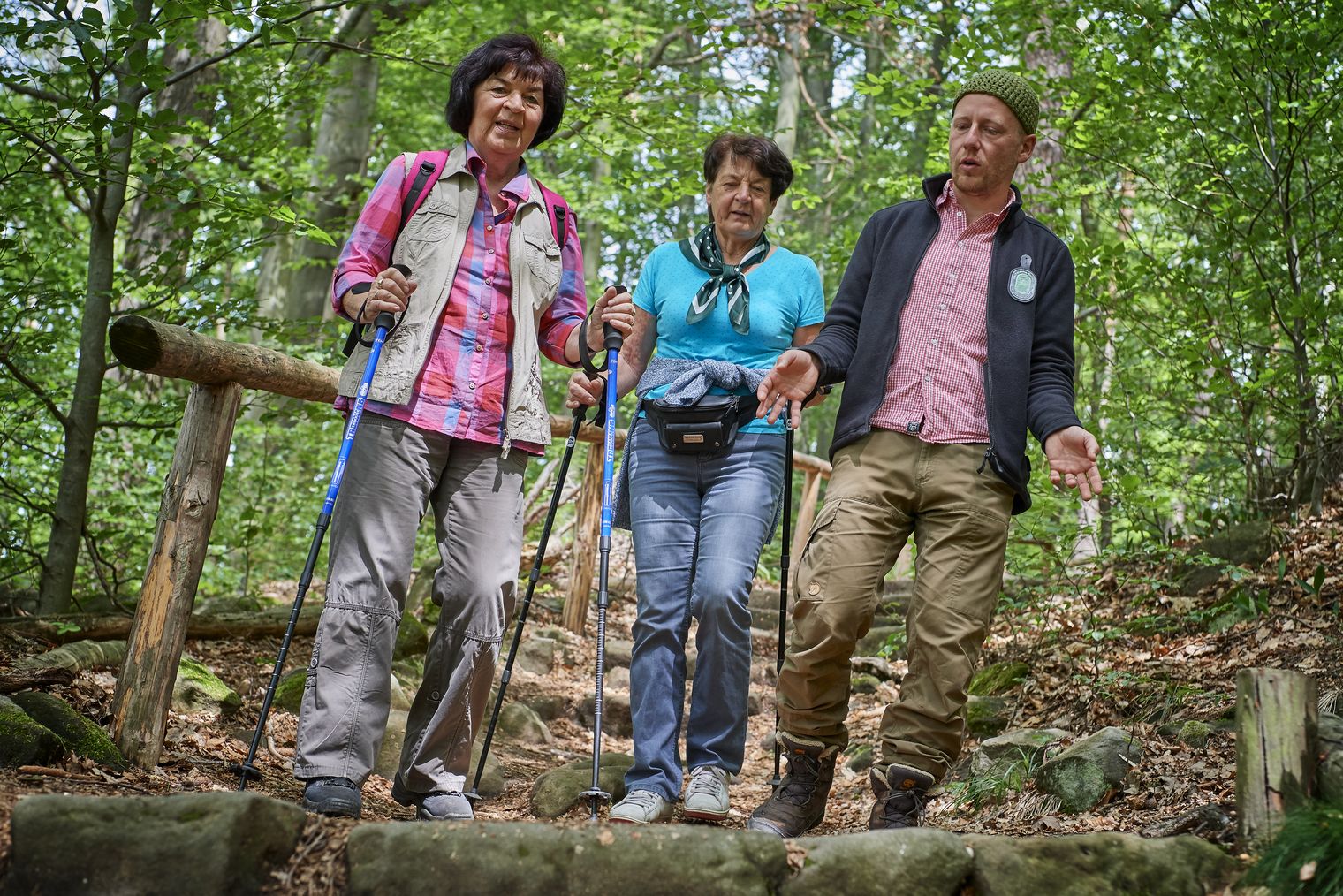Drei Personen wandern auf einem Waldweg, tragen Wanderstöcke und lächeln.