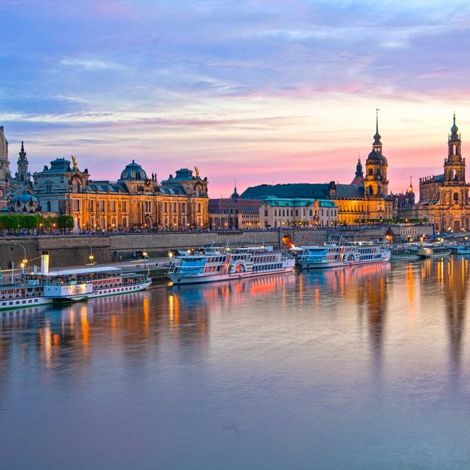 Dresden bei Sonnenuntergang, mit beleuchteten Gebäuden und Booten auf dem ruhigen Wasser.
