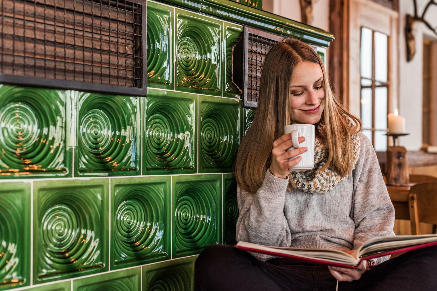 Frau mit Kaffeetasse sitzt am grünen Kachelofen der Wachbergbaude und liest ein Buch.
