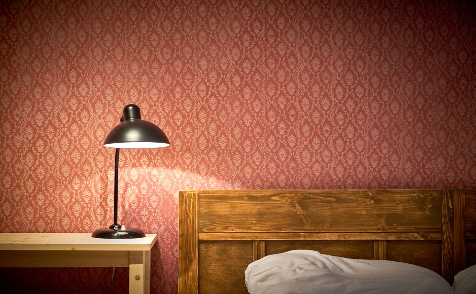 Holzbettrahmen neben einem Nachttisch mit einer schwarzen Tischlampe vor einer rot gemusterten Wand.