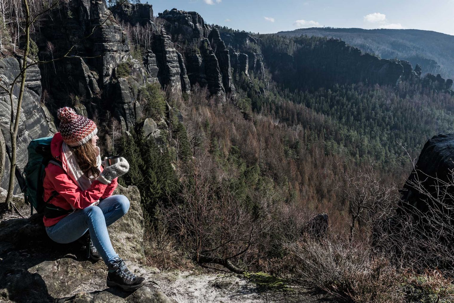 Frau in roter Jacke sitzt auf einem Felsen und genießt die Aussicht auf eine bewaldete Landschaft.