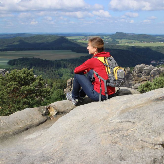 Person in roter Jacke sitzt auf einem Felsen und blickt auf eine grüne Landschaft mit Hügeln.