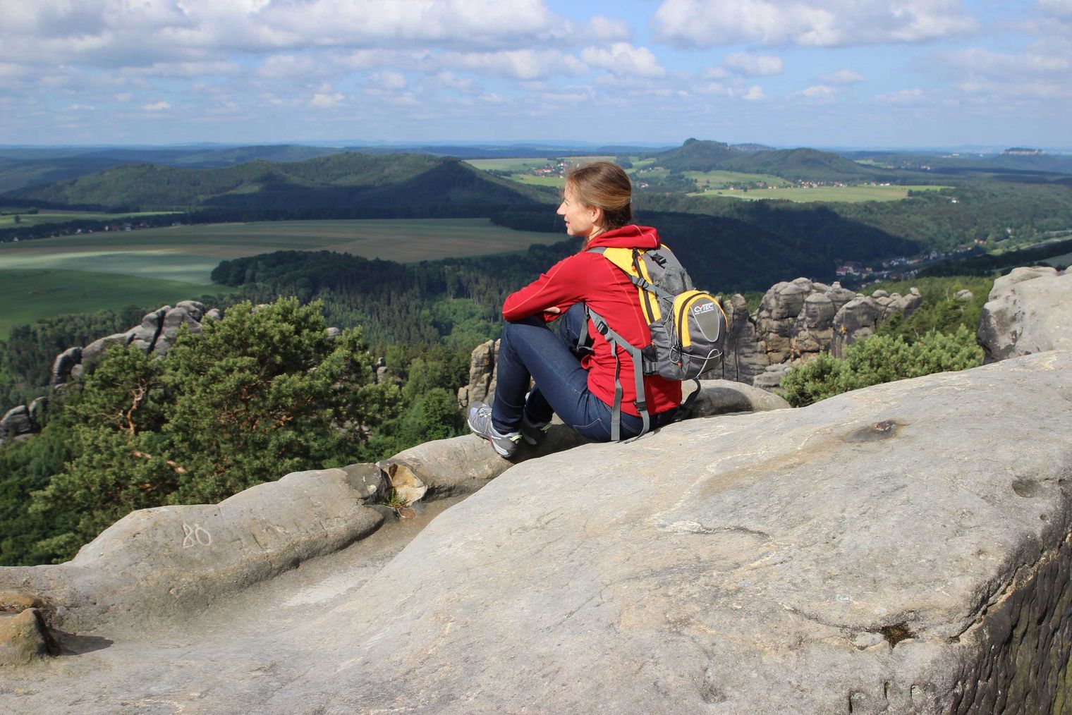 Person in roter Jacke sitzt auf einem Felsen und blickt auf eine grüne Landschaft mit Hügeln.