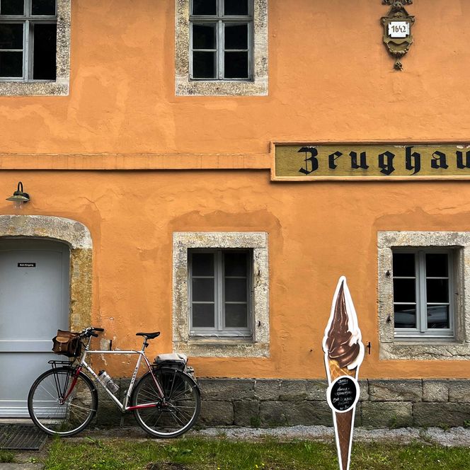 Orangefarbene Wand eines Gebäudes mit der Aufschrift "Zeughaus", daneben ein Fahrrad und eine Figur.