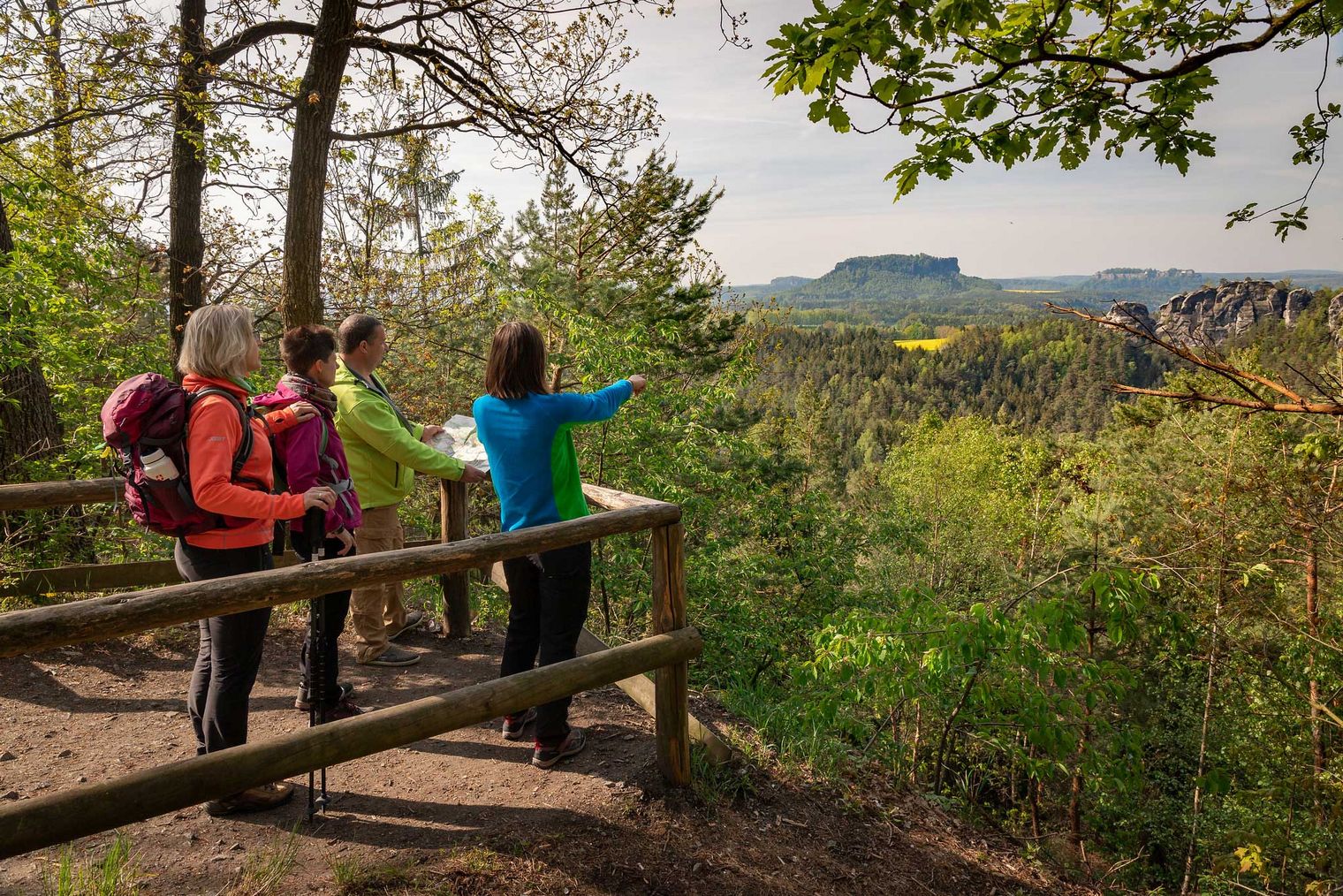 Drei Wanderer stehen an einem Aussichtspunkt und betrachten die Landschaft, umgeben von Bäumen.