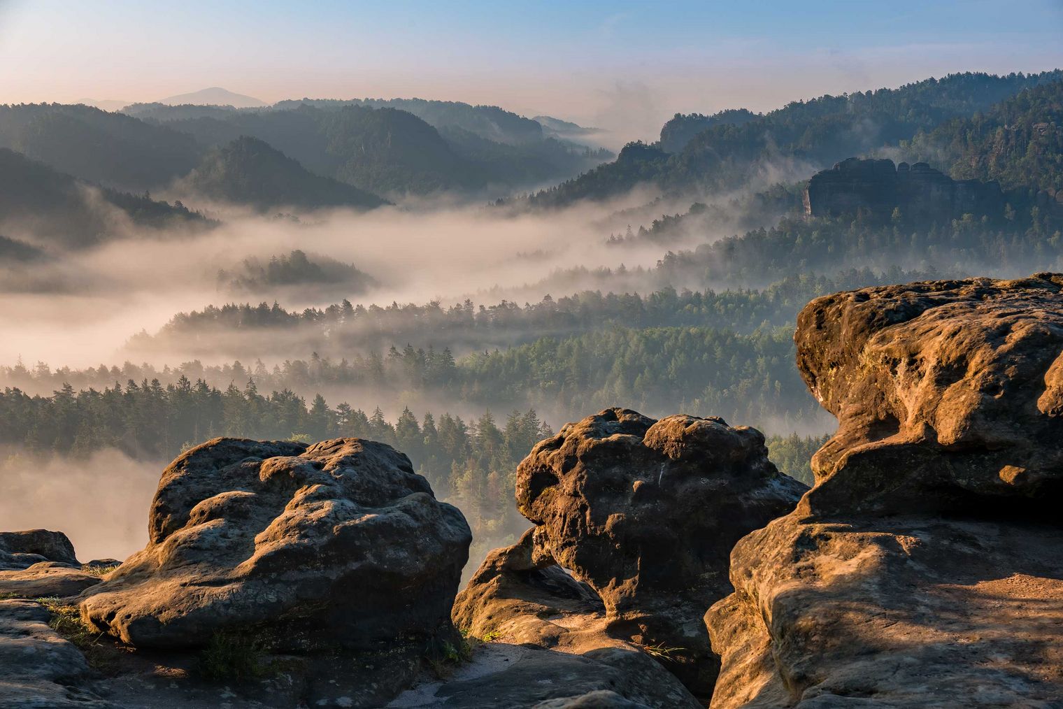 Blick auf eine neblige Landschaft mit Bergen und Wäldern im Hintergrund. Felsen im Vordergrund.