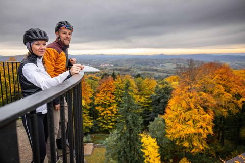 Paar mit Fahrradhelm an der Aussichtsterrasse des Unger Turm, umgeben von bunten Herbstbäumen und bewölktem Himmel.