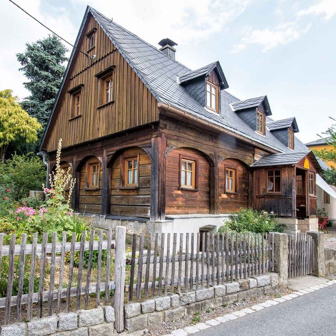 Holzhaus mit Satteldach, umgeben von Bäumen und einem Garten, eingezäunt mit einem Holzlattenzaun.