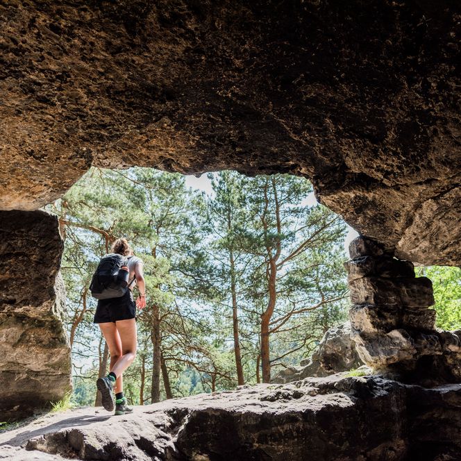Wanderer mit Rucksack steht in der Kleinsteinhöhle und blickt auf einen Wald mit Bäumen im Hintergrund.