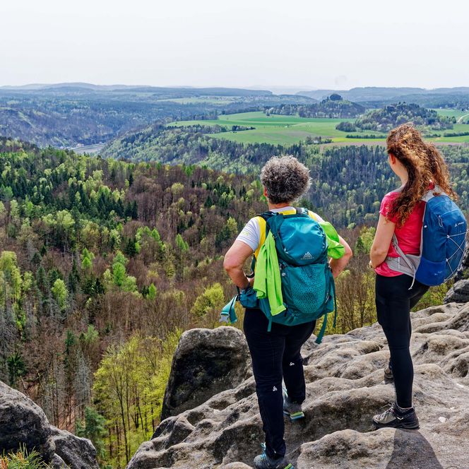Zwei Wanderer mit Rucksack stehen auf einem Felsen und blicken auf eine grüne Landschaft.