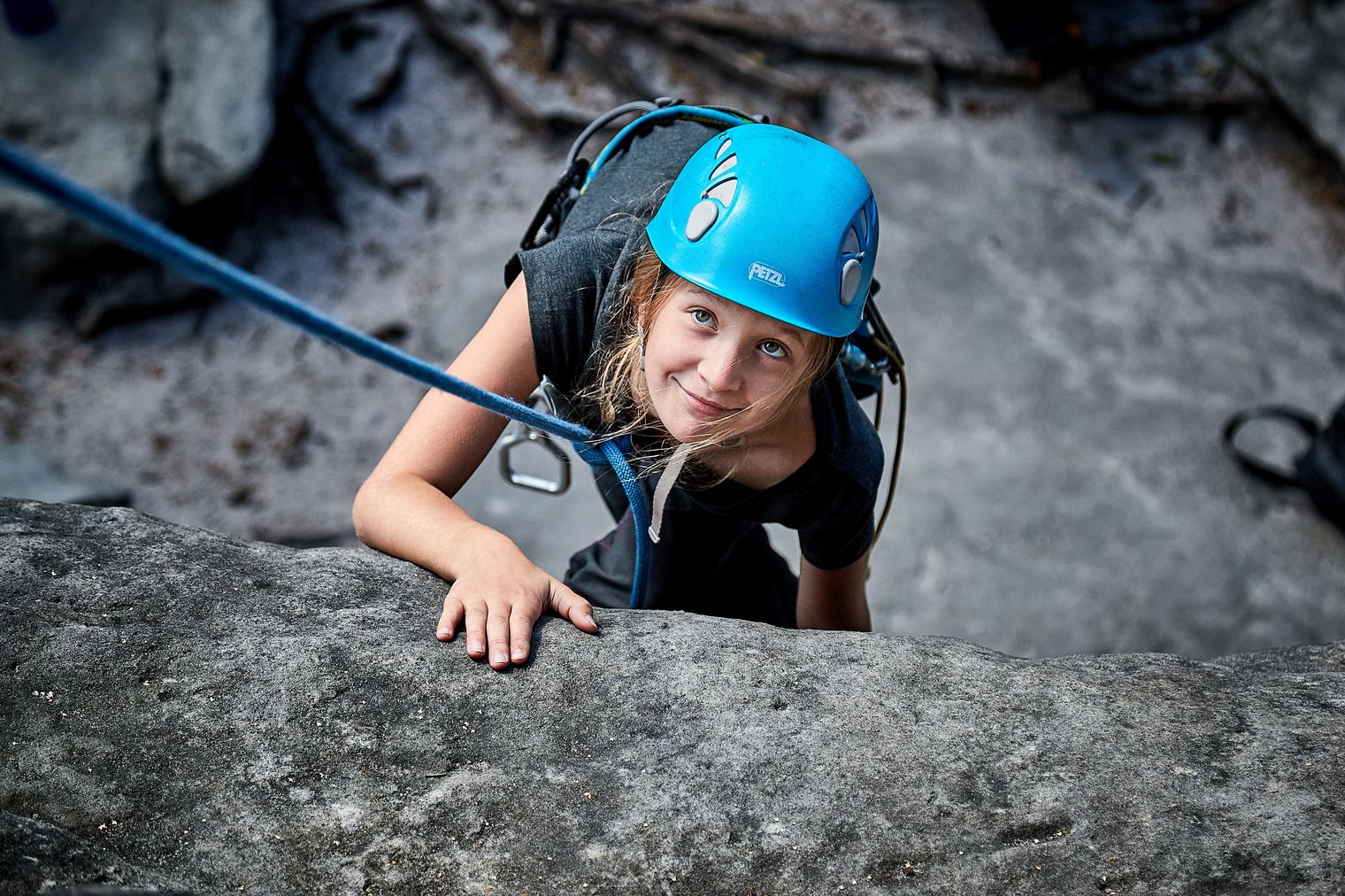 Kind mit blauer Kletterhelm klettert an einer Felswand, schaut nach oben.