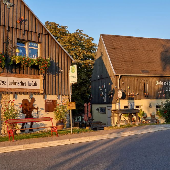 Zwei rustikale Gebäude mit Holzfassade, umgeben von Pflanzen und Sitzgelegenheiten im Freien.
