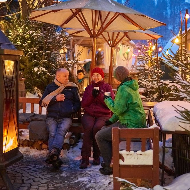 Drei Personen sitzen unter einem Sonnenschirm in einer winterlichen Umgebung mit Schnee und Lichtern.