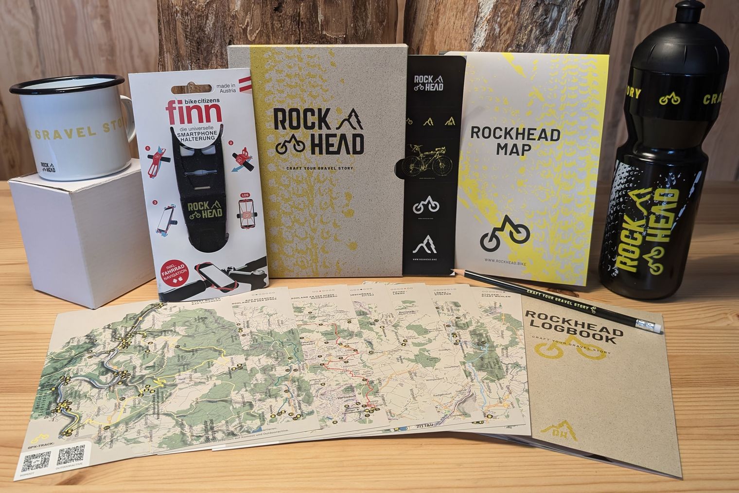 Starterpaket des RockHeads bestehend aus Karte, Trinkflasche, Emaille-Tasse und weiterem vor einem Holzuntergrund.