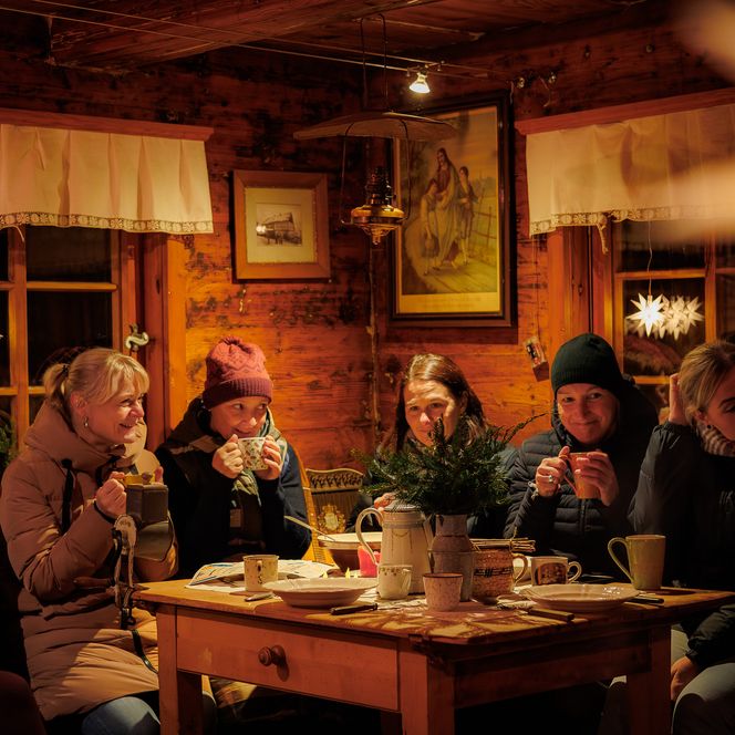 Gemütliche Runde von Menschen in einem Holzraum, die an einem Tisch sitzen und Getränke halten.