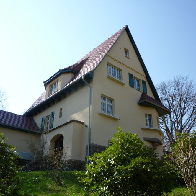 Zweistöckiges, gelbes Haus mit rotem Satteldach und grünen Fensterläden, umgeben von Bäumen und Sträuchern.