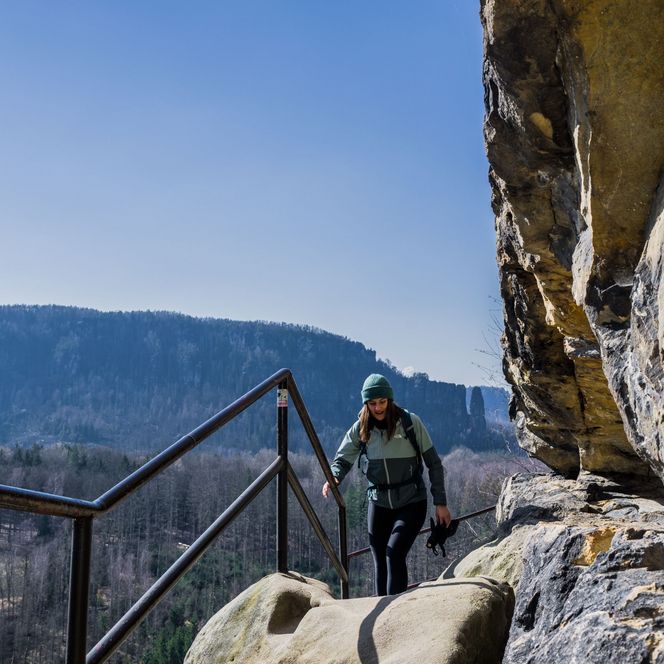 Wanderer steigt eine Treppe entlang einer Felswand in einer bergigen Landschaft.