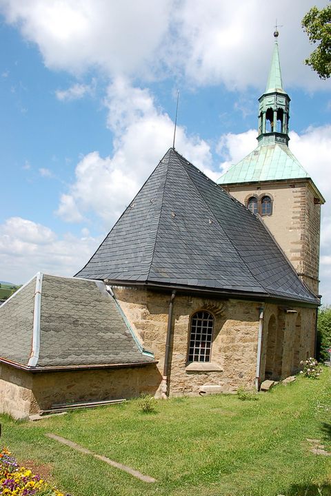 Kirche mit dunklem Dach, umgeben von Bäumen, blauer Himmel im Hintergrund.