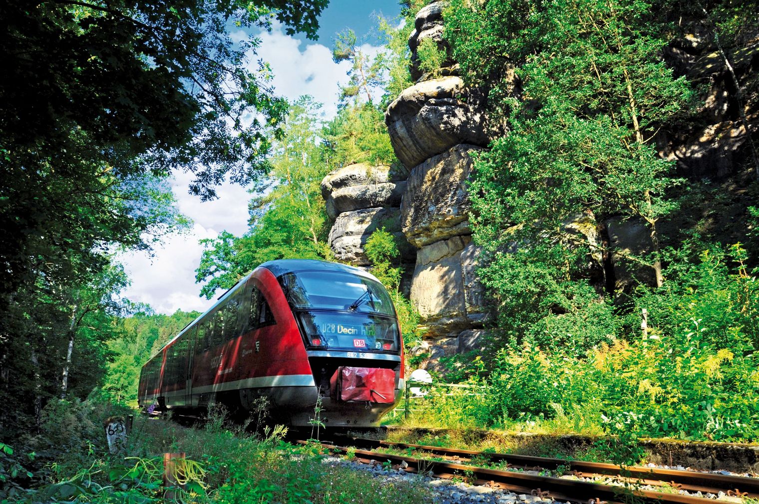 Roter Zug fährt auf einer Schiene durch eine grüne Landschaft mit Felsen im Hintergrund.