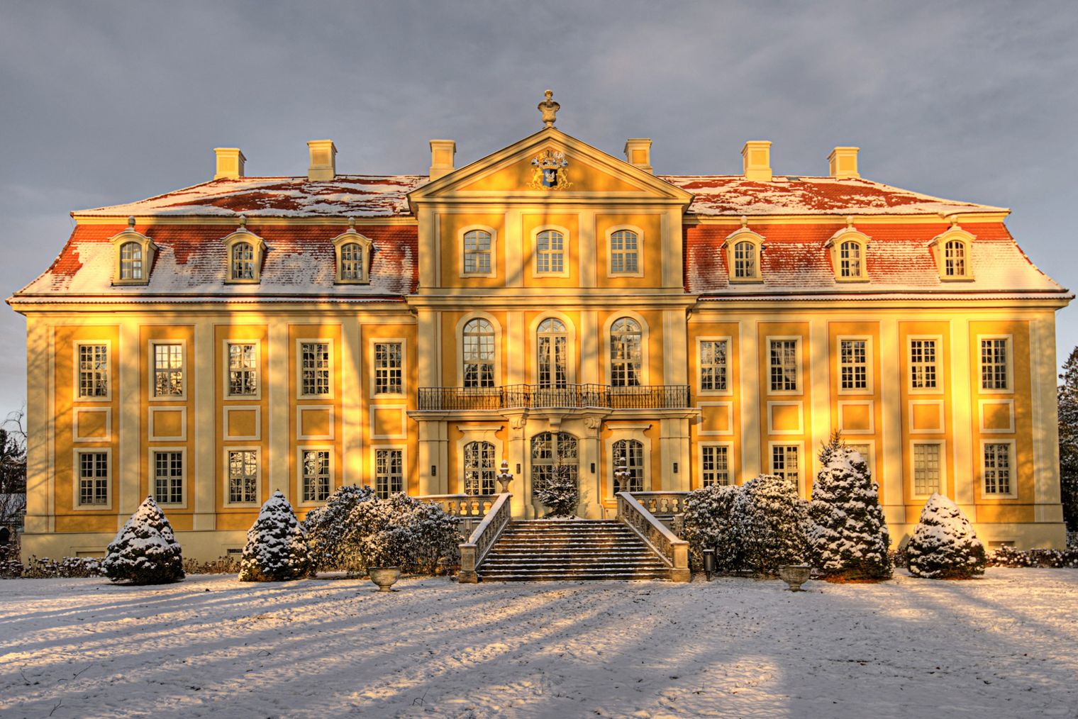 Historisches Gebäude mit gelber Fassade, Schnee auf dem Boden und Bäumen, im warmen Licht der Abenddämmerung.