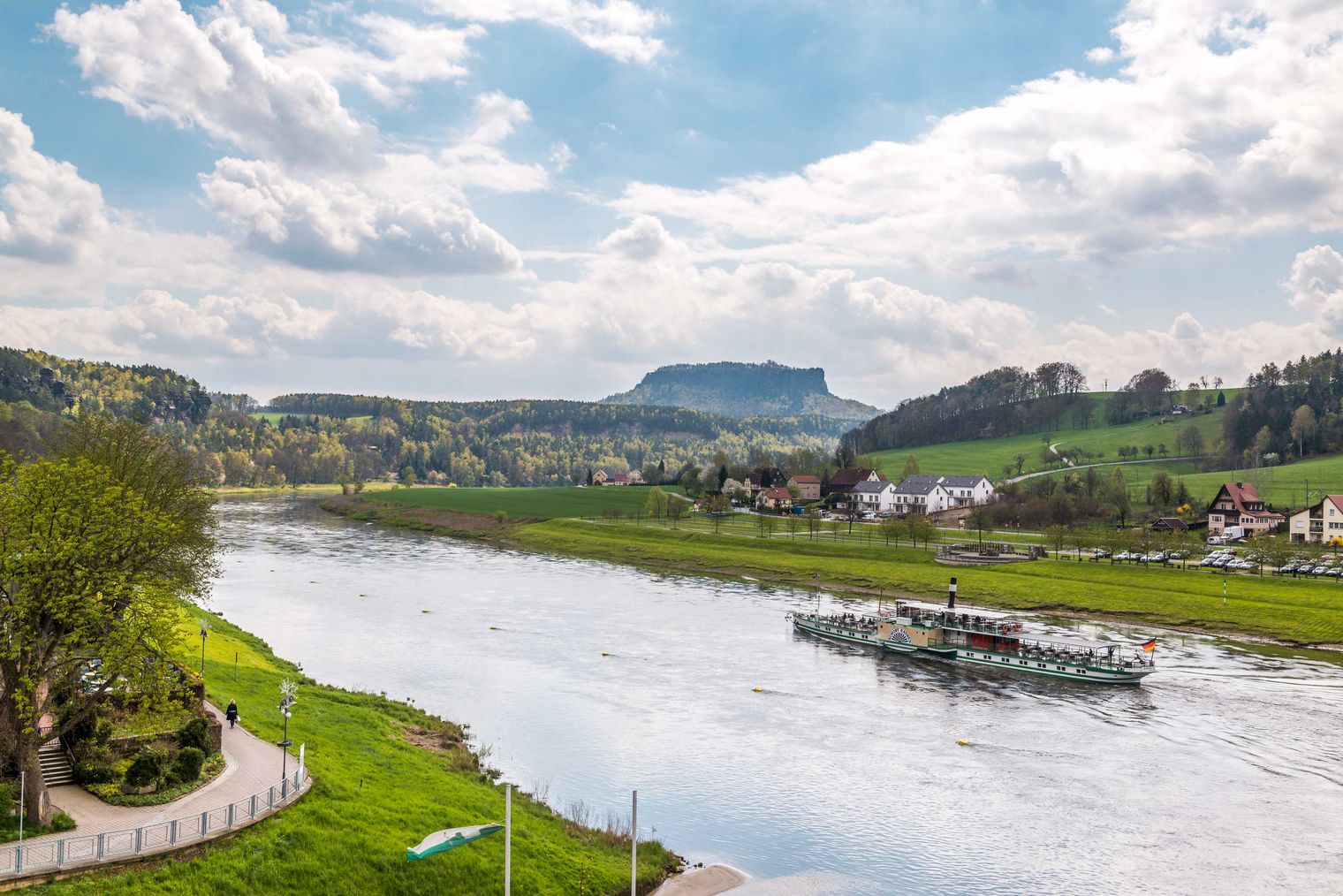 Blick auf einen Fluss mit grünen Ufern und bewaldeten Hügeln im Hintergrund unter einem bewölkten Himmel.