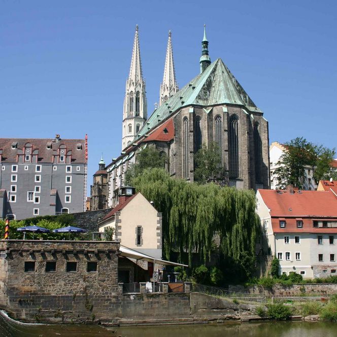 Historisches Gebäude mit zwei spitzen Türmen, umgeben von Bäumen und Wasser.