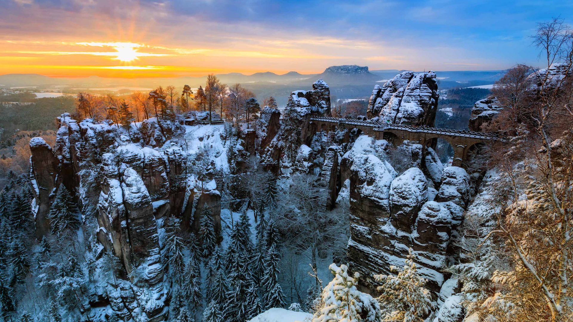 Schneebedeckte Felsenlandschaft mit einer Brücke, Sonnenaufgang im Hintergrund.